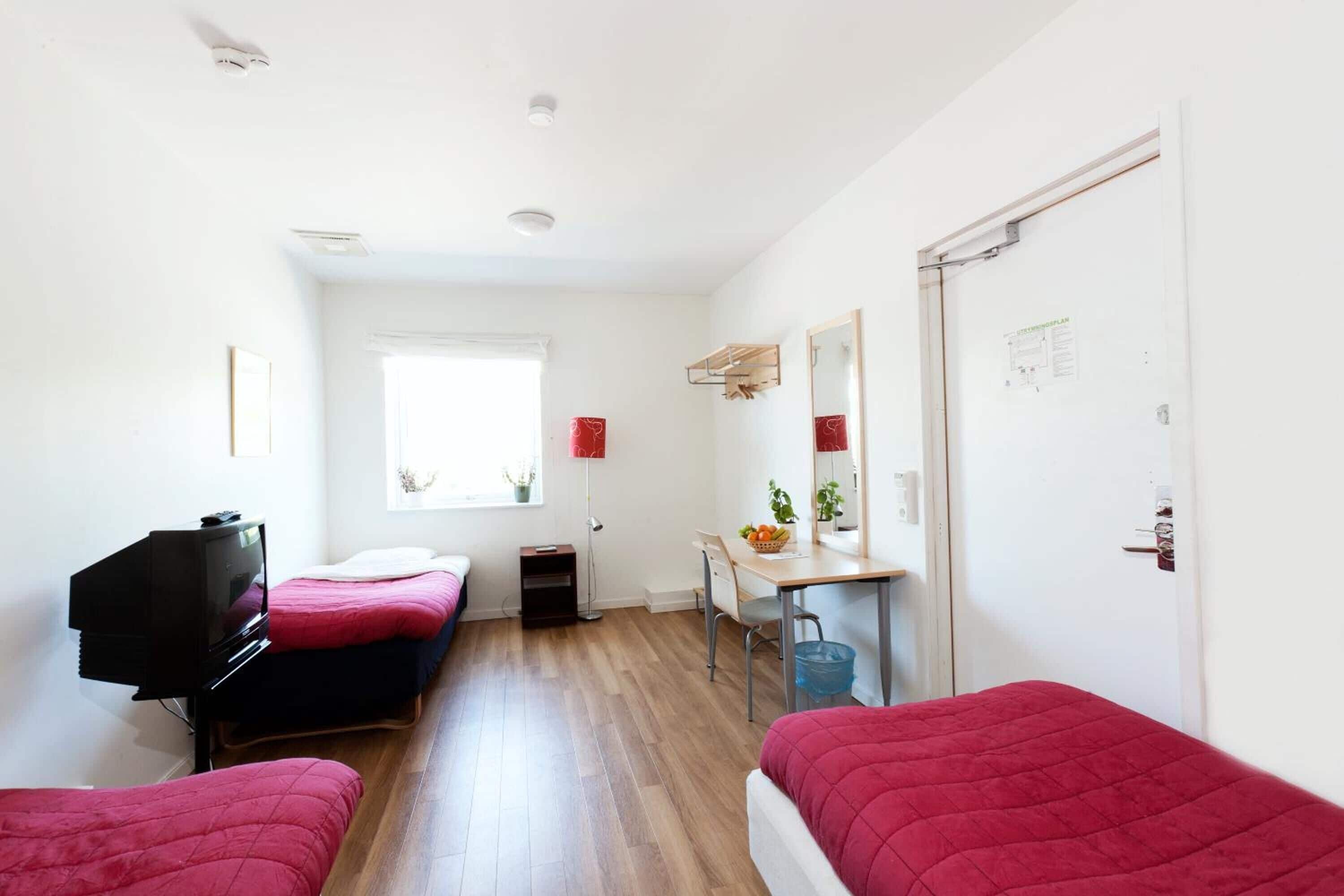 Una habitación pequeña y luminosa con dos camas, un escritorio y un televisor.