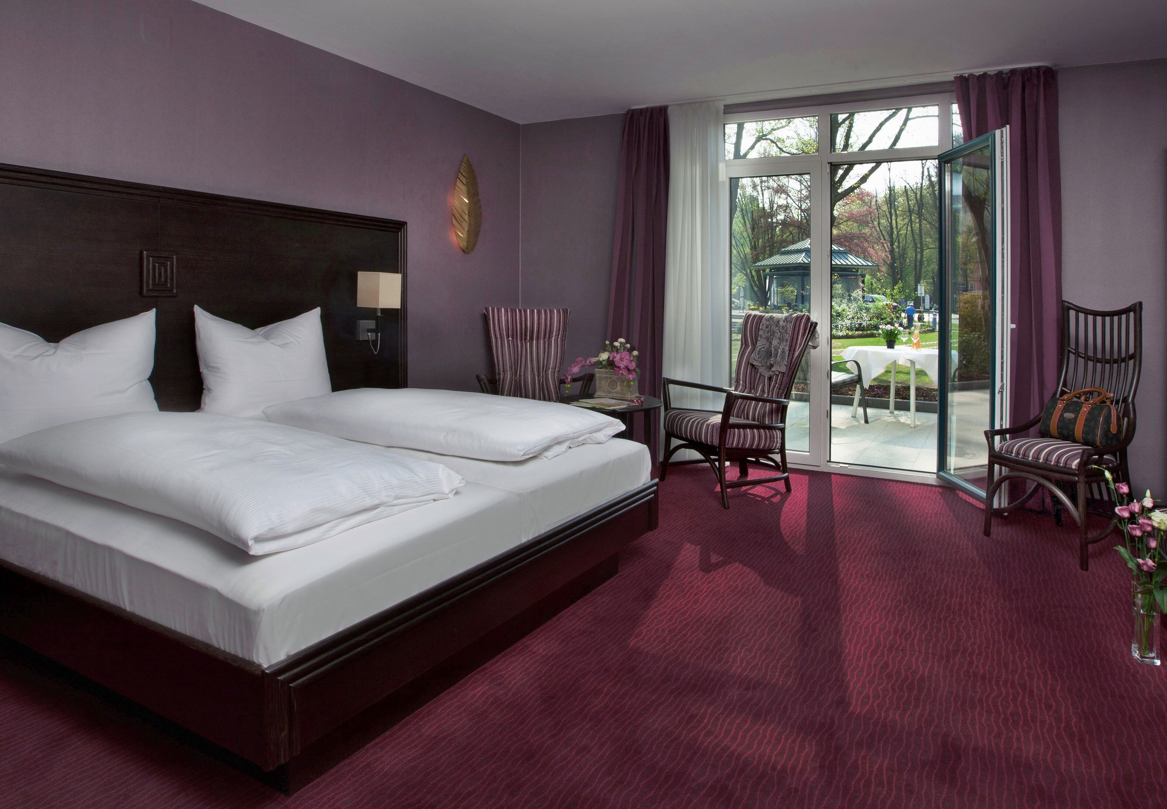 Ein Hotelzimmer mit einem großen Bett, zwei Sesseln, einem Tisch und Blick auf den Garten durch eine Glastür.