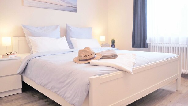 Cama bem arrumada com roupa de cama branca e um chapéu de palha, uma mesa de cabeceira com copos e um livro, e um quadro pendurado na parede.