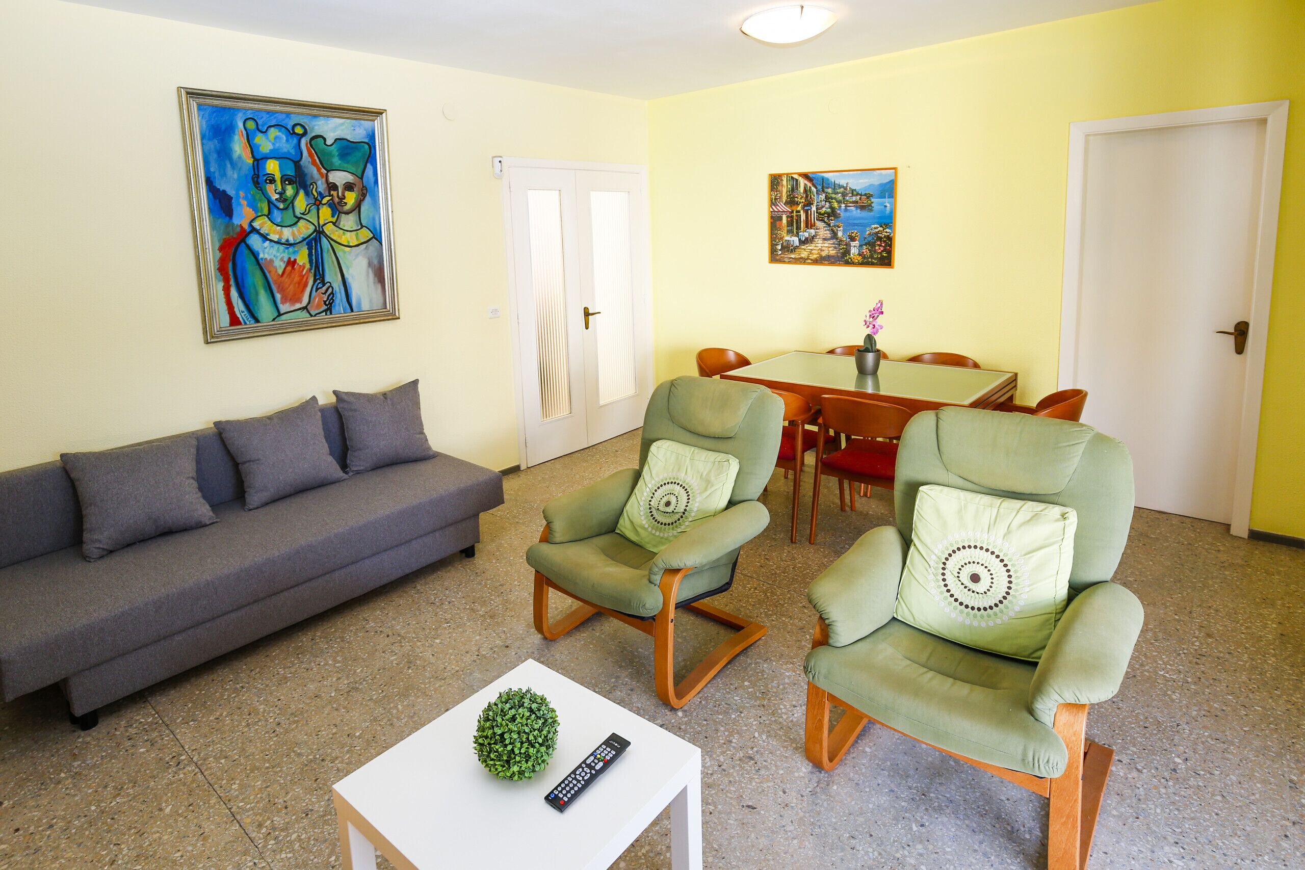 Un salon comprenant un canapé, deux fauteuils, une table basse et une table à manger avec des chaises. Des tableaux ornent les murs, et une porte est visible au fond.