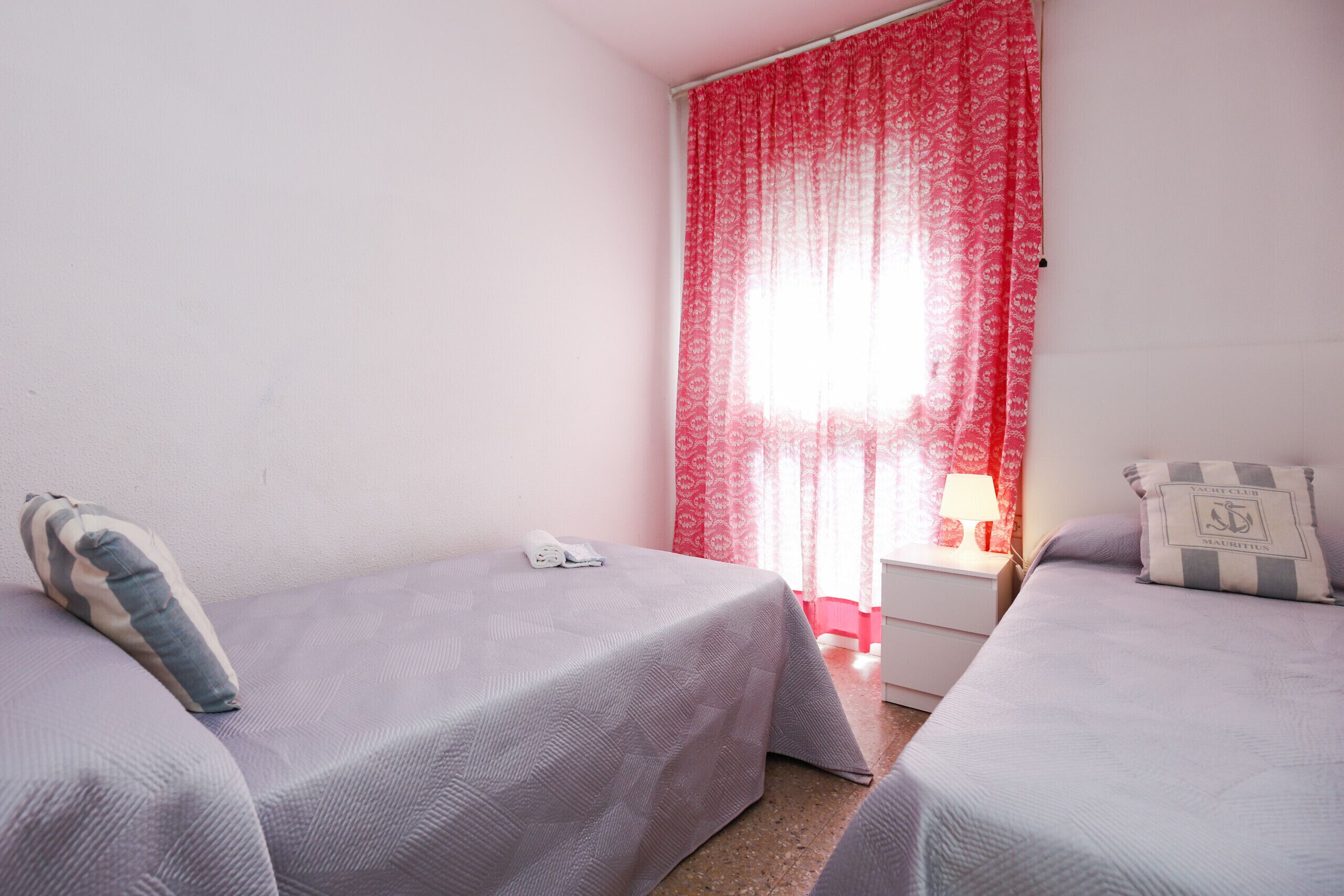 Una habitación con dos camas, una mesita de noche, una lámpara y una ventana con cortinas rojas.