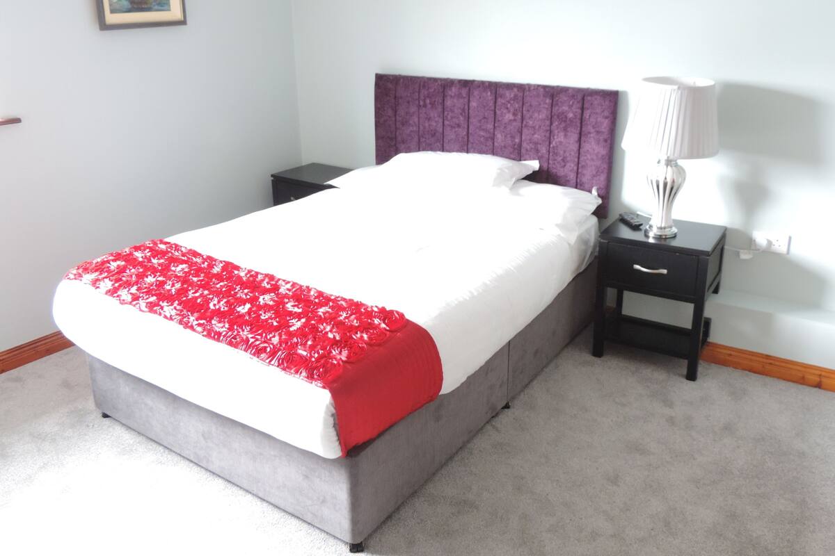 Un lit bien fait, recouvert d’une couvre-lit blanche et rouge, avec une tête de lit violette et une table de chevet surmontée d’une lampe.