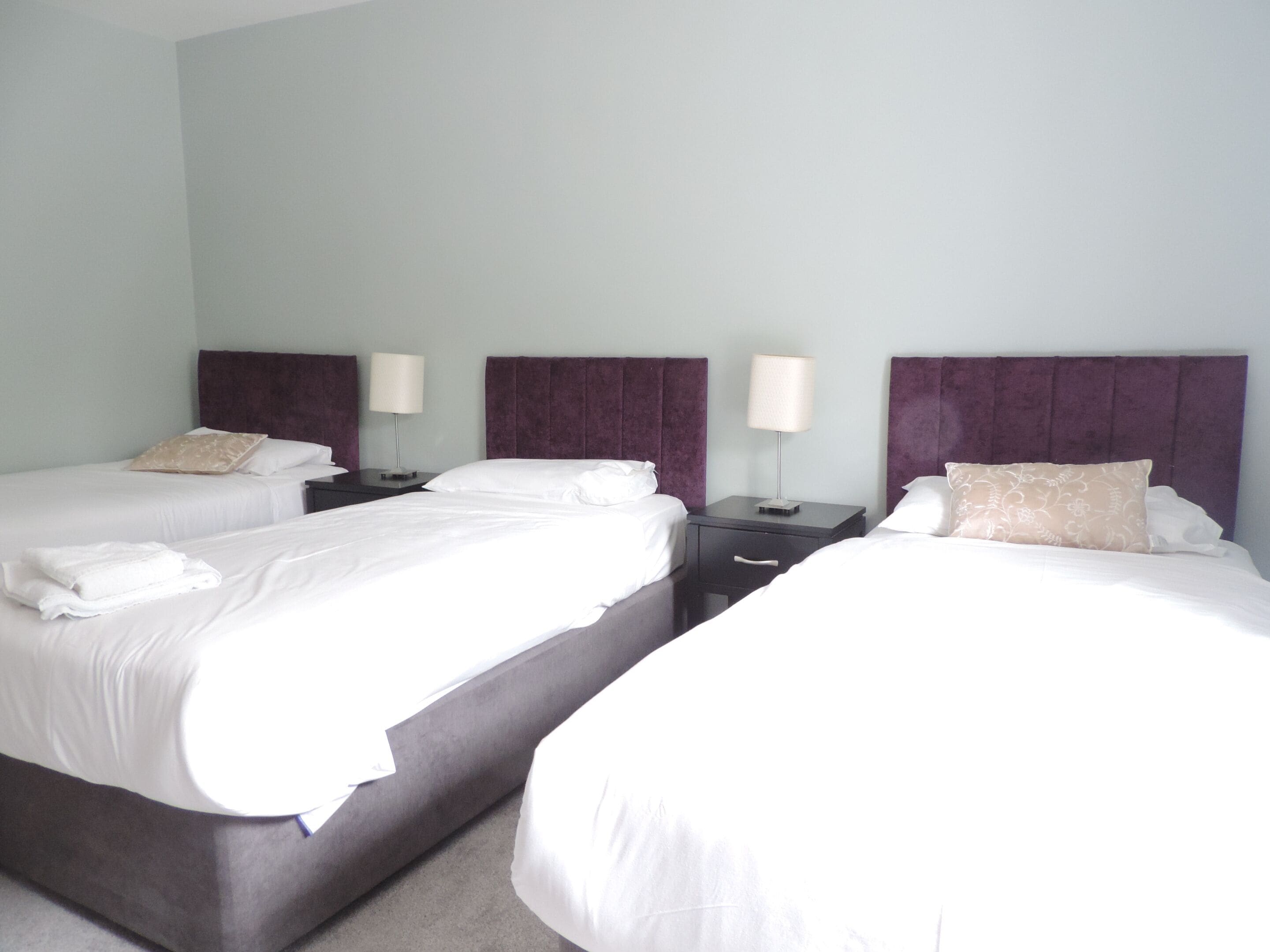A hotel room with two beds, each with white linens and a headboard. There are two bedside tables with lamps and a small drawer.