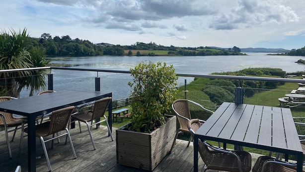 An outdoor deck with tables and chairs overlooking a river, surrounded by greenery and hills.