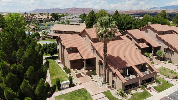A residential area with modern houses featuring red-tiled roofs, a central courtyard with a palm tree, and a view of mountains in the background. - NEWLY UPGRADED Beautiful One Bedroom, One Bath, Sports Village Condo (St. George)