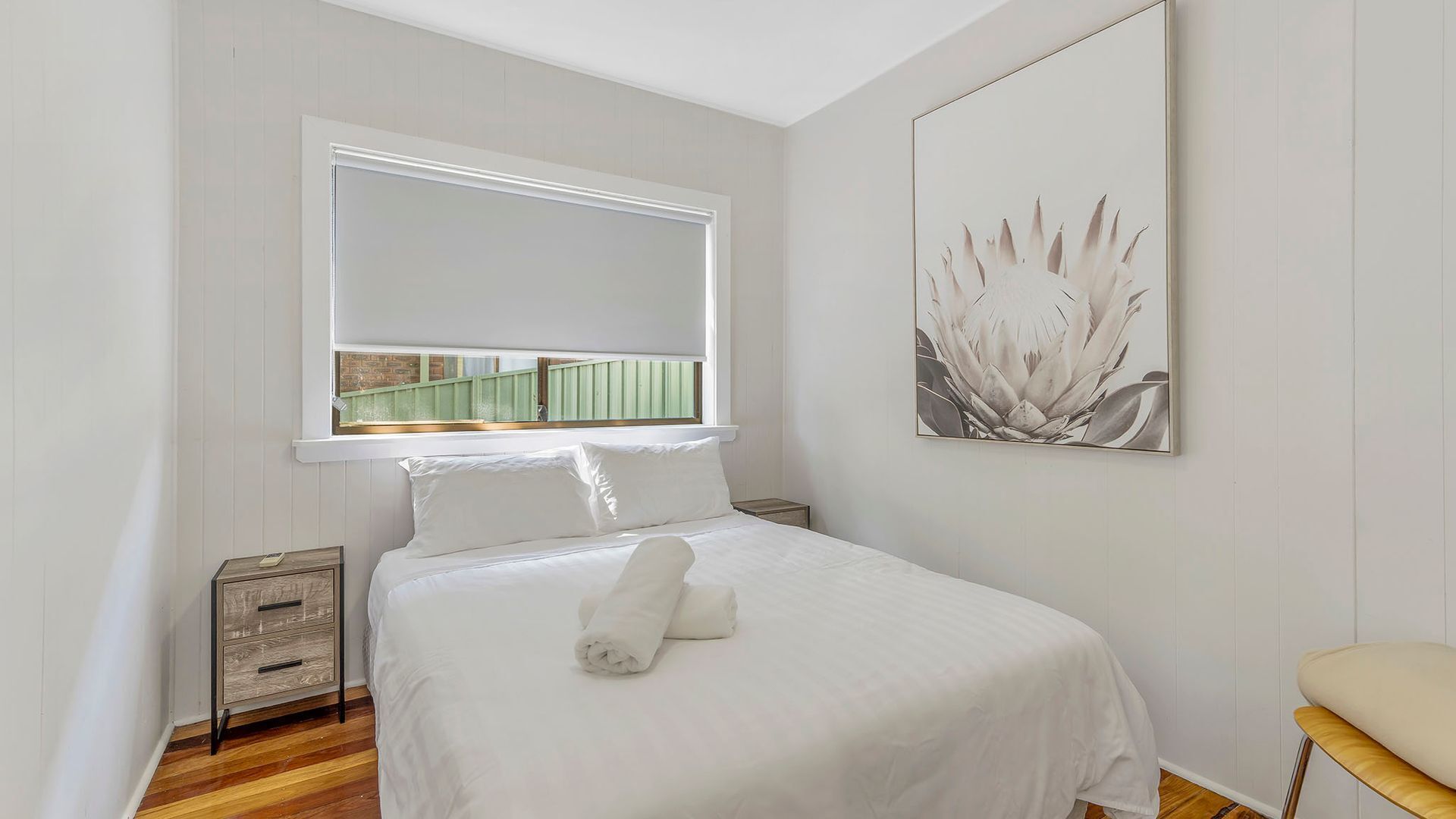 A bedroom with a bed, a window, a nightstand, and a wall-mounted artwork.