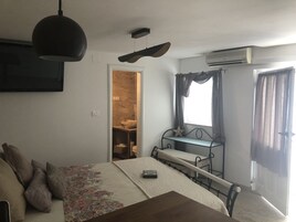 A bedroom with a bed, bedside table, and a small desk with a chair. There is a window with curtains, an air conditioning unit, and a door leading to a bathroom.