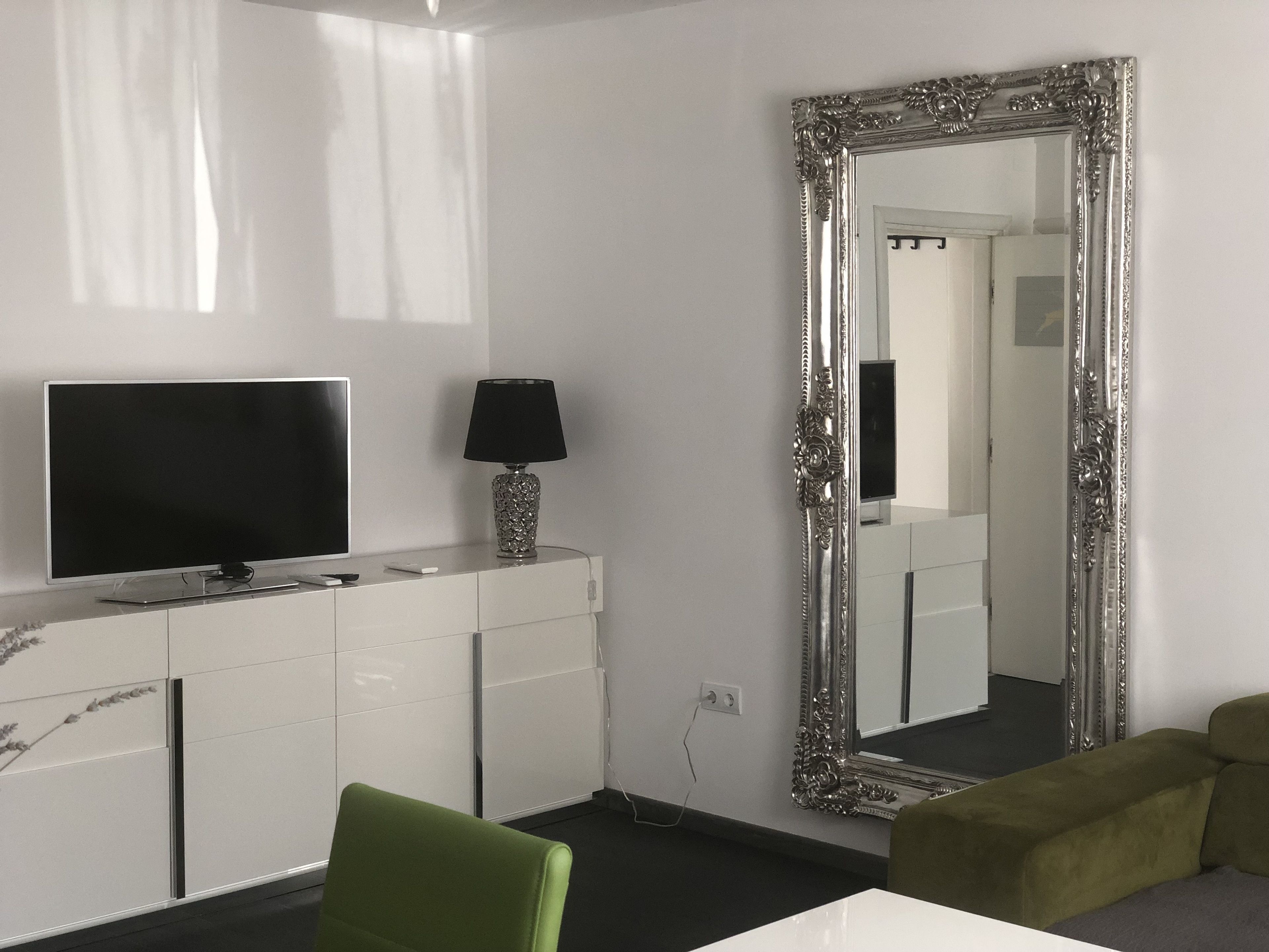 A modern hotel room with a large mirror, a flat-screen TV, a lamp, and a green chair.