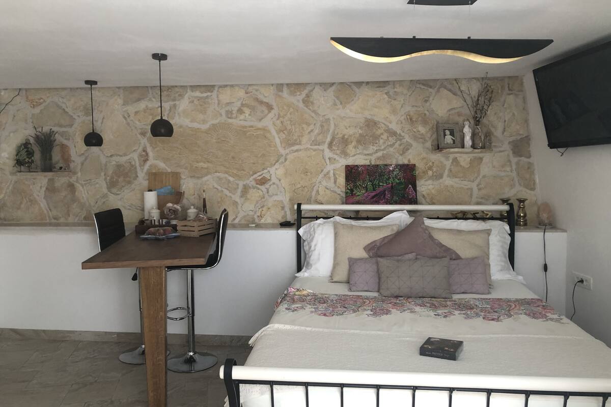 Une chambre avec un mur en pierre, un lit avec de nombreux oreillers, une table de chevet, un bureau avec une chaise, un luminaire suspendu et un téléviseur fixé au mur.