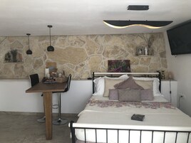 Un dormitorio con pared de piedra, una cama con muchos cojines, una mesita de noche, un escritorio con una silla, una lámpara colgante y un televisor montado.