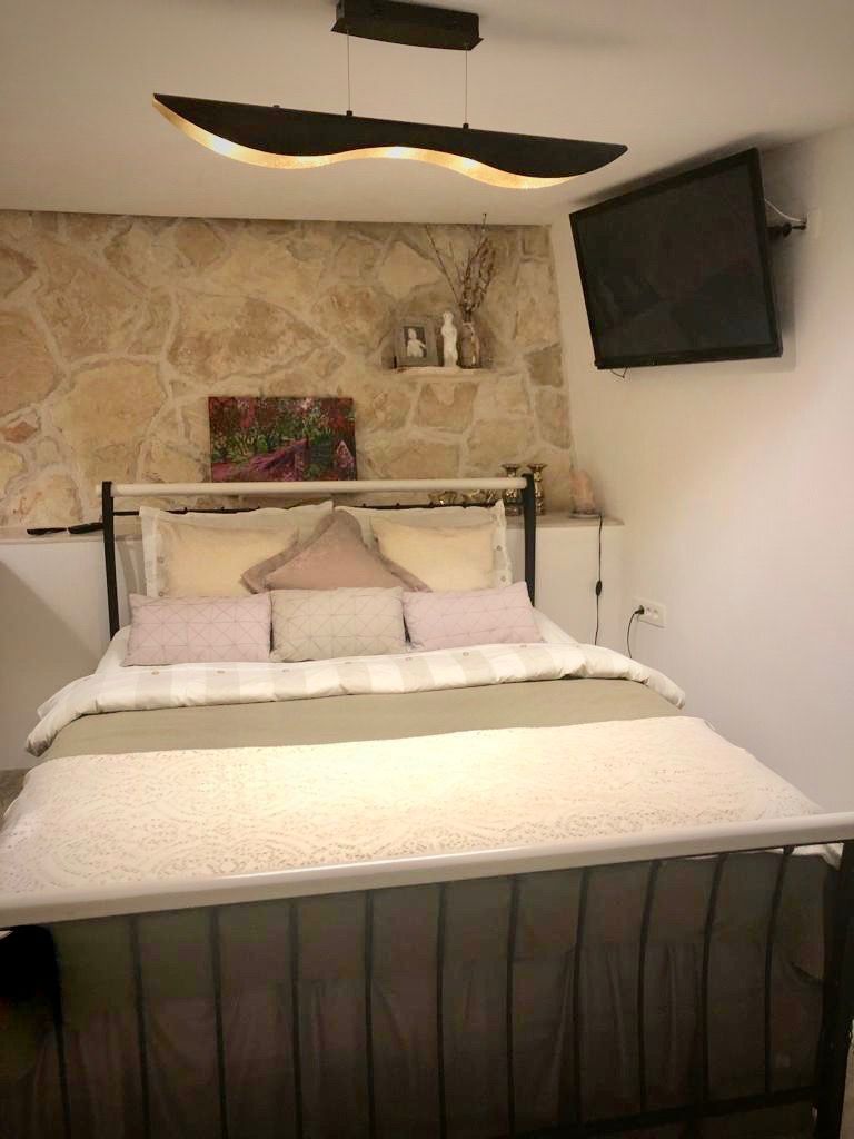 A bed with a headboard against a stone wall, a flat-screen TV mounted on the wall, and a pendant light fixture above the bed.