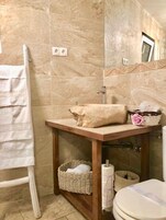 Un baño con pared de mármol, tocador de madera, lavabo blanco, perchero, cesta de mimbre y un inodoro.