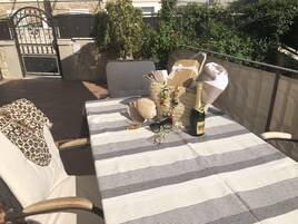Mesa al aire libre con mantel a rayas, una botella de champán, gafas de sol, un bolso tejido y objetos decorativos.