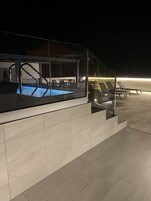 Un área de piscina moderna al aire libre con cerramiento de vidrio, sillas de descanso y una escalera que sube a un nivel superior.