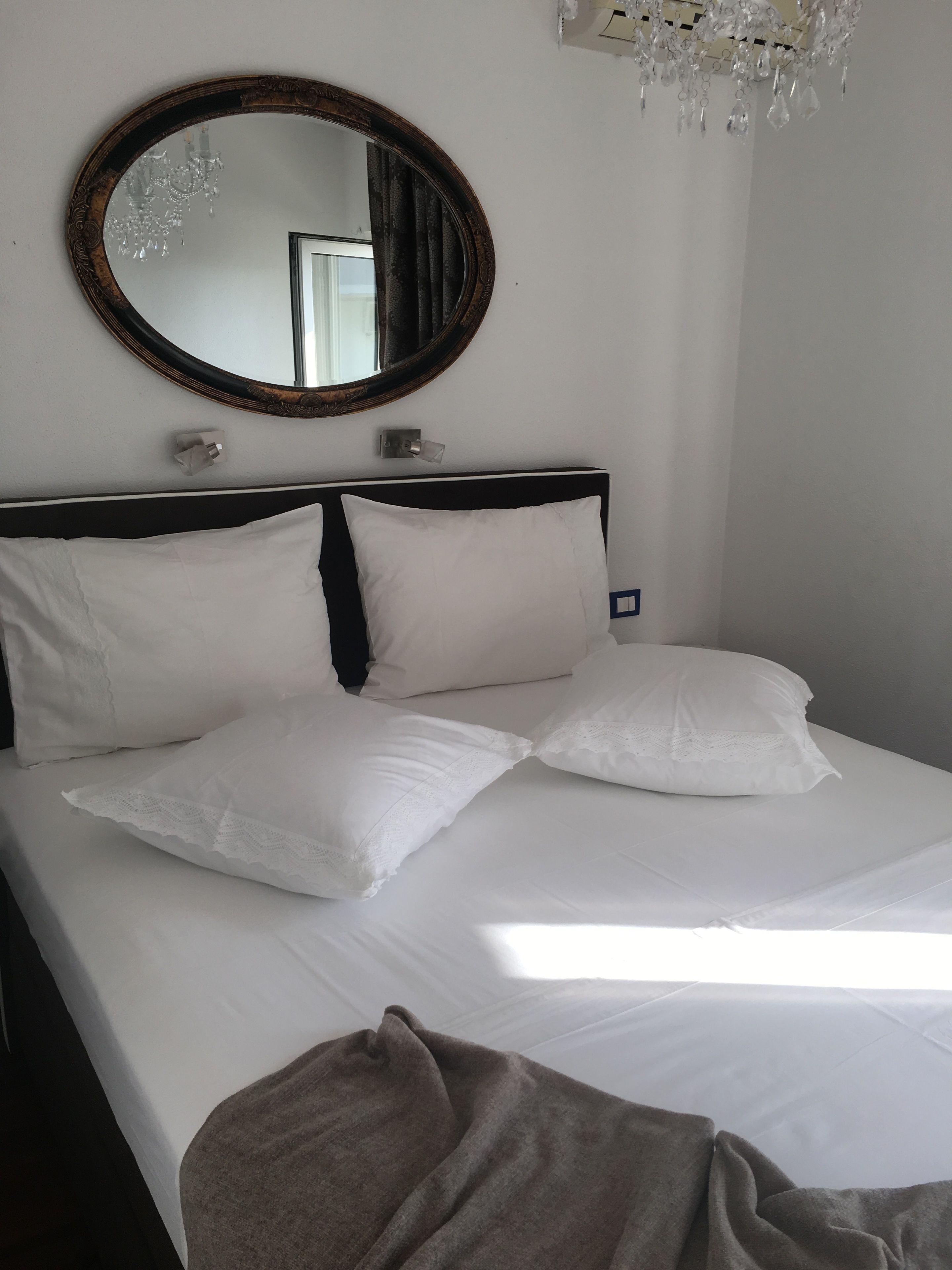 Un lit bien fait, avec du linge de lit blanc et une tête de lit sombre. Un miroir rond, encadré de bois foncé, est accroché au-dessus du lit.