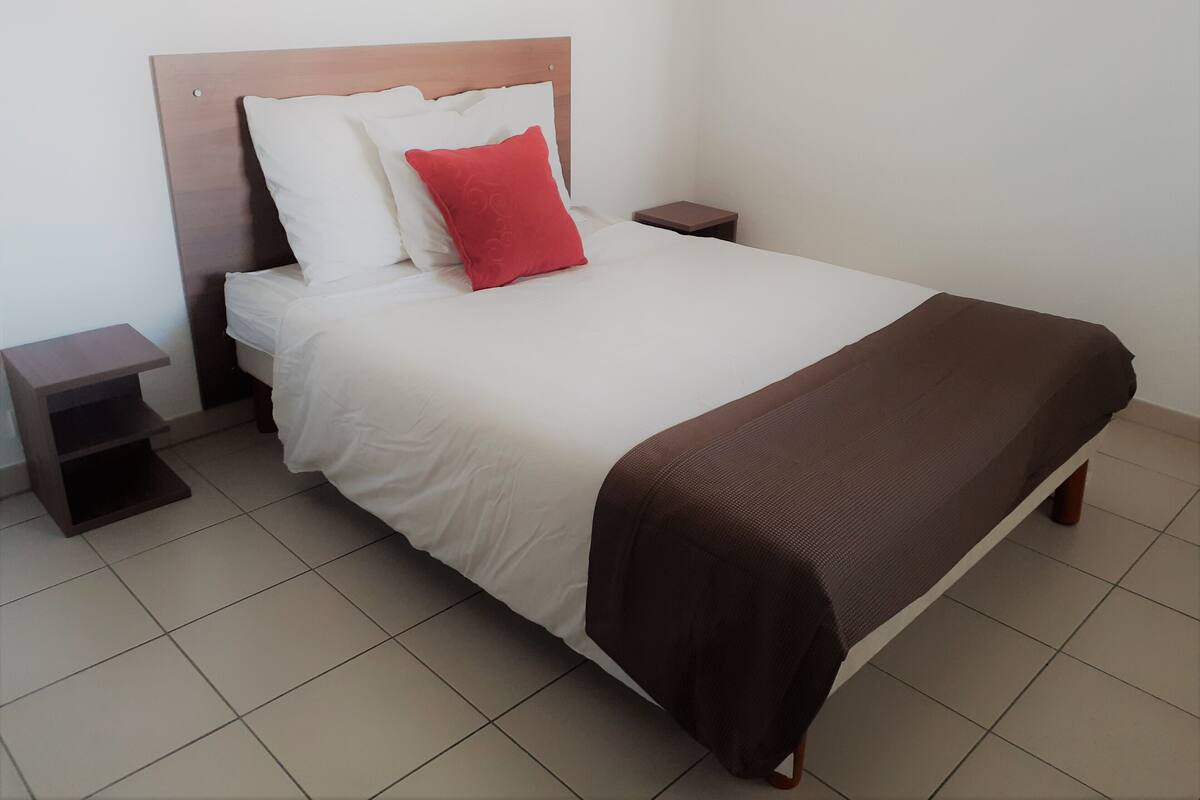 Een net opgemaakt bed met een houten hoofdbord, witte en rode kussens, een bruine deken en twee nachtkastjes.