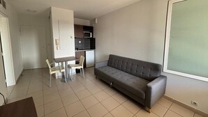 A living room with a sofa, a dining table with chairs, a kitchenette, and a large window. - Garden & City Toulon (Toulon)