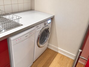 Eine kompakte, weiße Waschmaschine mit Frontlader, einer Bedienblende und einer Edelstahl-Tür, platziert in einer Ecke eines Raumes mit Holzboden und einer gefliesten Rückwand.