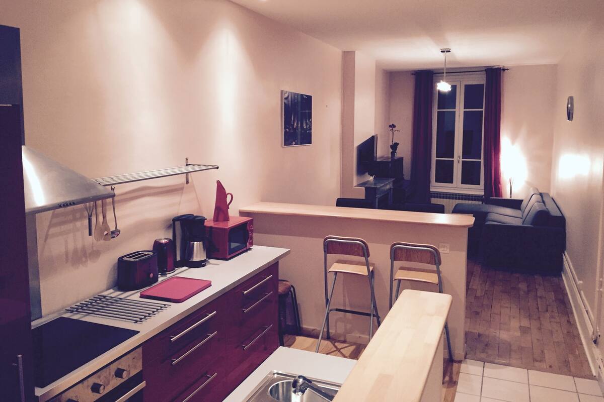Eine kompakte Küche mit Mikrowelle, Toaster und Wasserkocher. Zwei Stühle und ein kleiner Esstisch sind vorhanden. Im Hintergrund ist ein Fenster mit Vorhängen zu sehen.