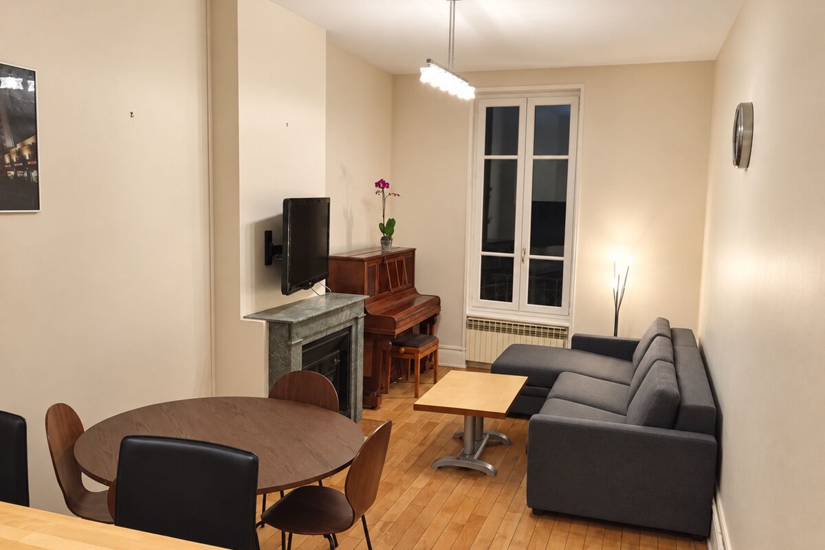 Ein modernes Wohnzimmer mit einem grauen Sofa, einem Holzesstisch mit Stühlen, einem an der Wand montierten Fernseher und einem Klavier.
