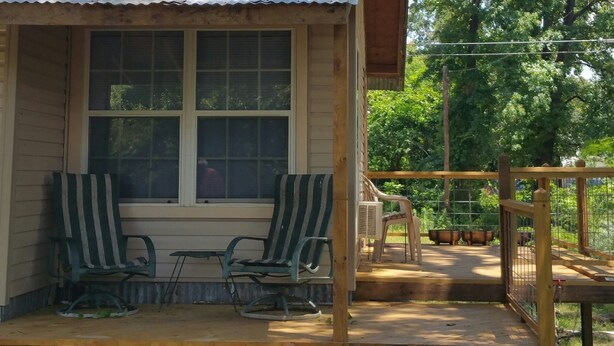 A wooden porch with two striped chairs and a table, a window, and a view of a fenced garden area. - Little lake house/ Fish cabin get away (Chandler)