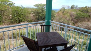 Un espacio al aire libre cubierto con una mesa y sillas, rodeado por una barandilla blanca, con vistas a un paisaje natural de árboles y arbustos.