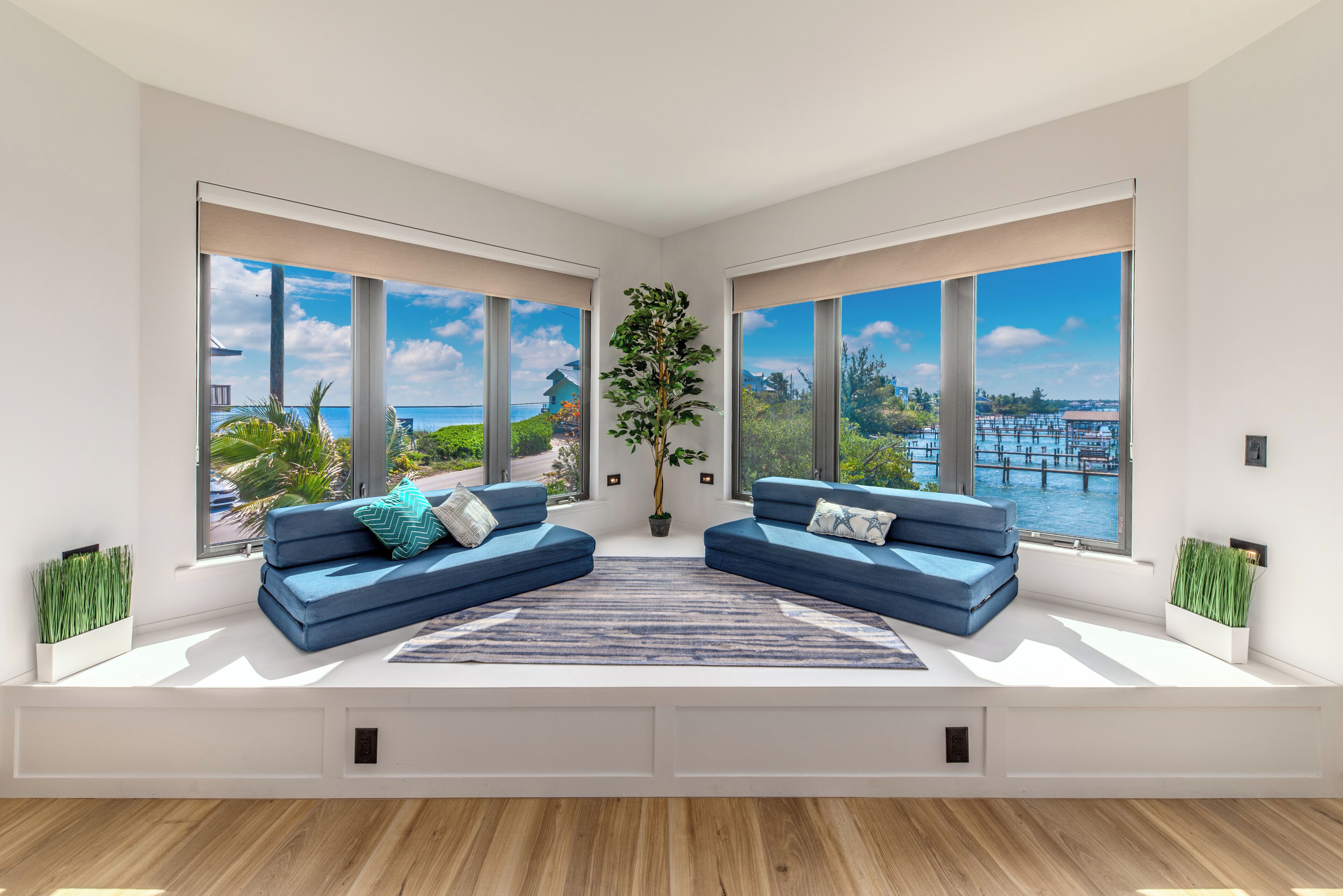 Een moderne woonkamer met grote ramen, een blauwe zithoek, een potplant en uitzicht op een waterkant.