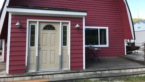A red barn-style building with a beige front door, two windows, and a small patio area with a table and chairs. - Furnished studio apartment (Unalaska)