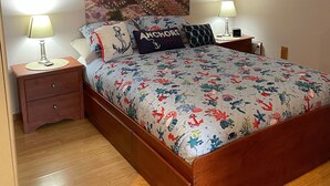 A bedroom with a wooden bed, two bedside tables with lamps, a large floral-patterned bedspread, and wall art depicting underwater scenes and a crab. - Furnished studio apartment (Unalaska)