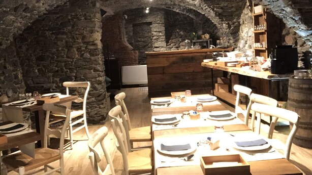 A rustic stone-walled dining area with wooden tables and chairs, a bar area with shelves, and a stone fireplace in the background. - NicoMat Rooms (Domodossola)