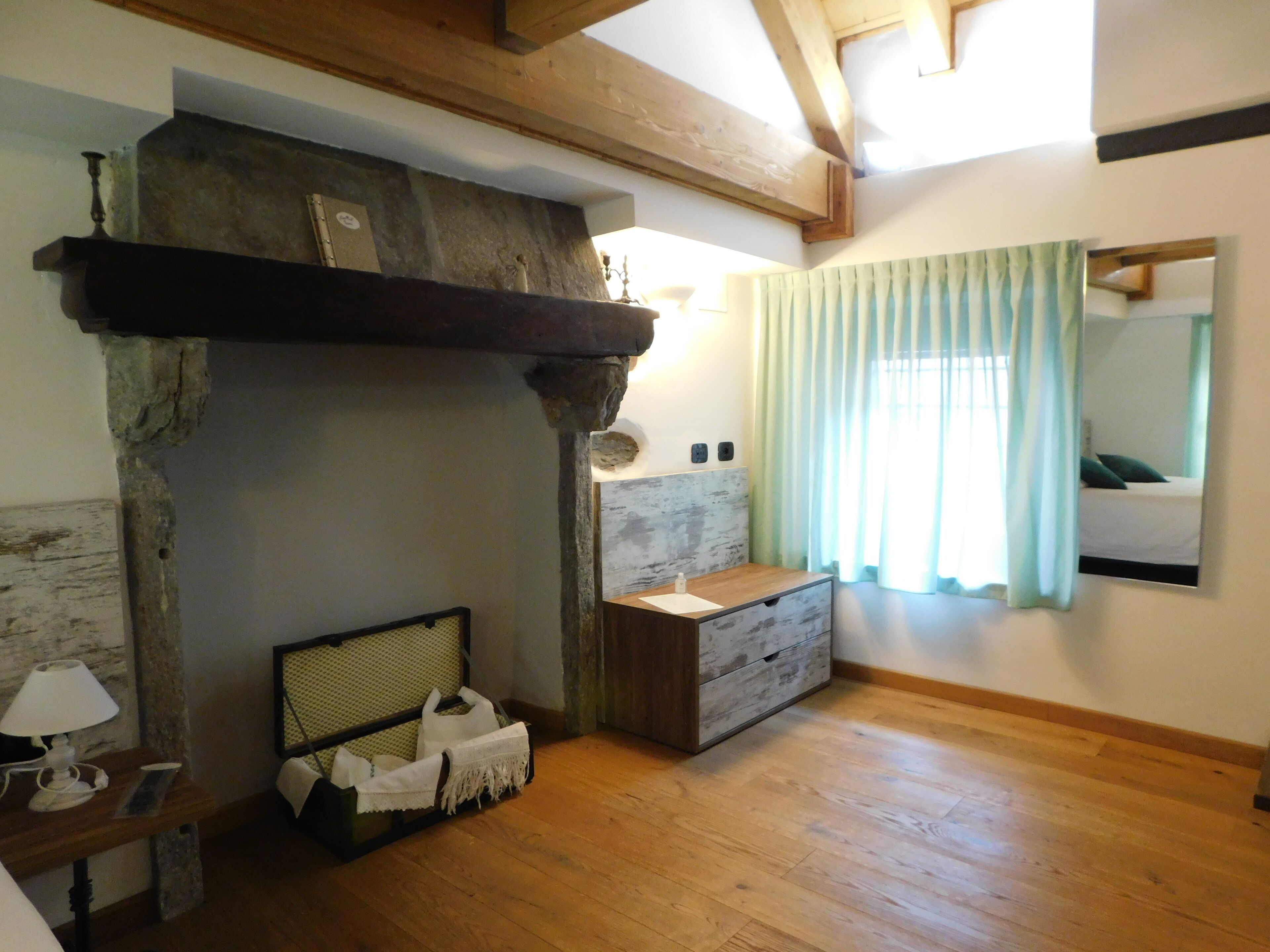 A room with a stone fireplace, wooden beams, a bed with a green curtain, and a bedside table with a lamp.