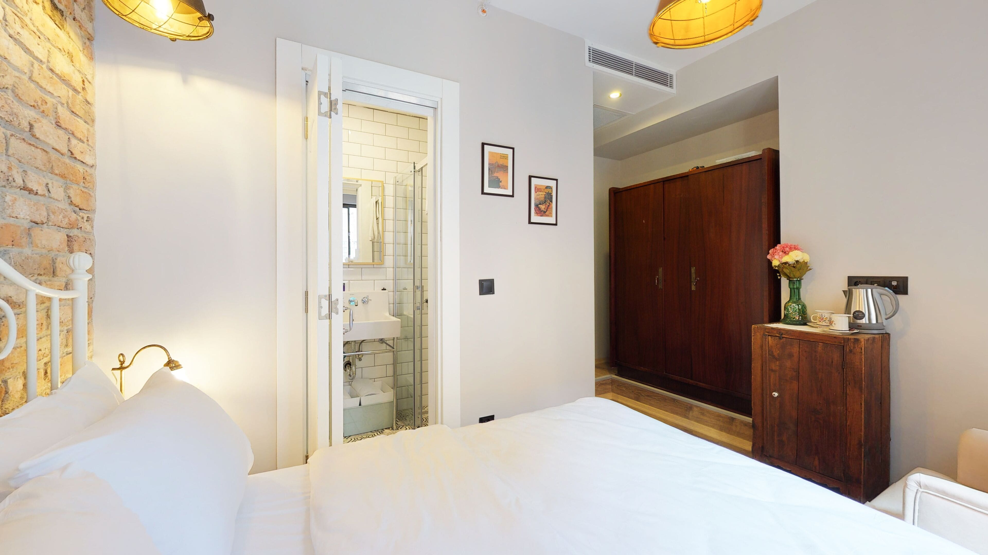 A bedroom with a bed, a wooden wardrobe, a small cabinet with a vase, and a door leading to a bathroom.