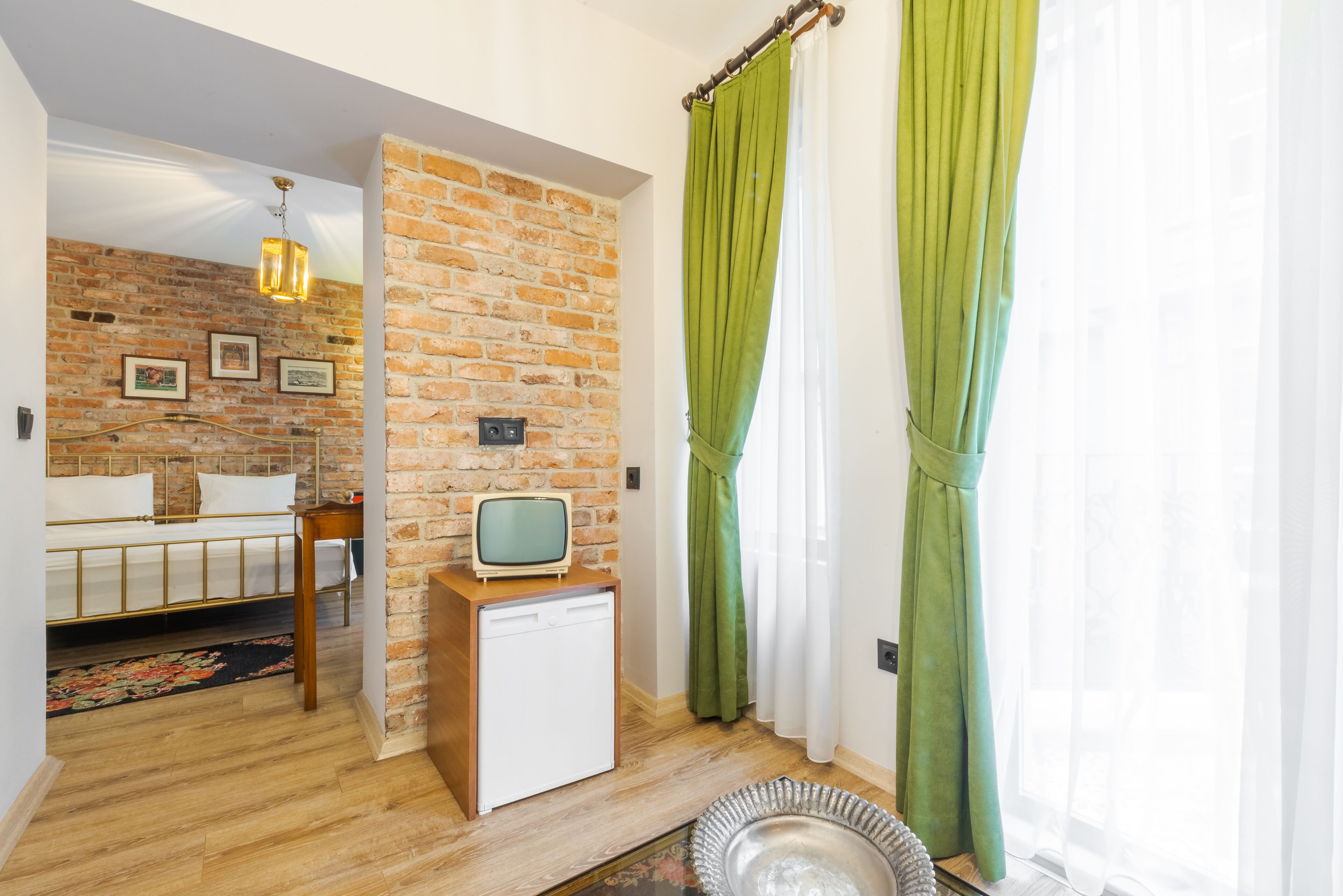 Une pièce avec un mur en briques, un téléviseur ancien posé sur un meuble en bois, un lit avec une literie blanche et un rideau vert.