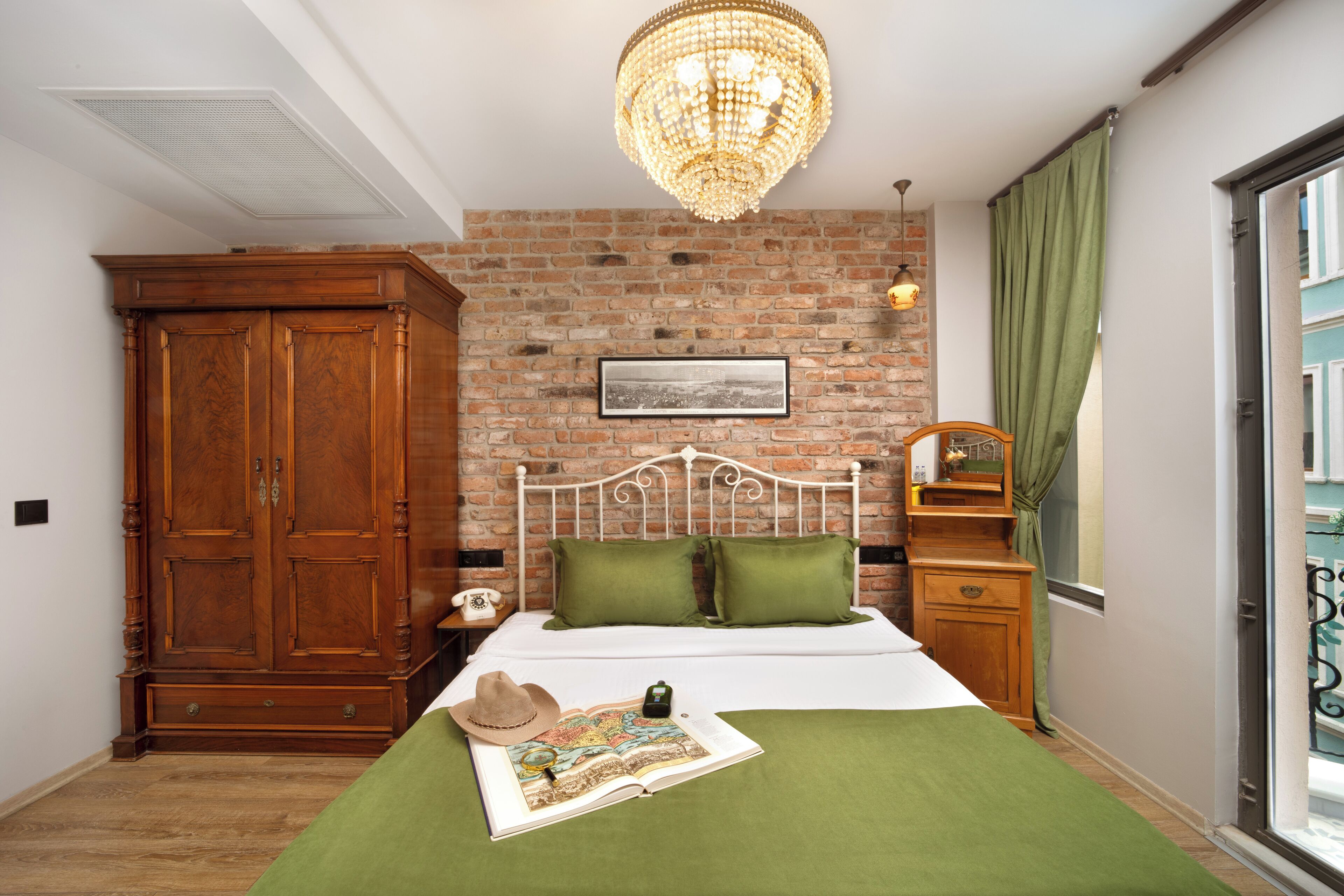 A bedroom with a brick wall, a wooden wardrobe, a bed with green bedding, a chandelier, and a window with curtains.