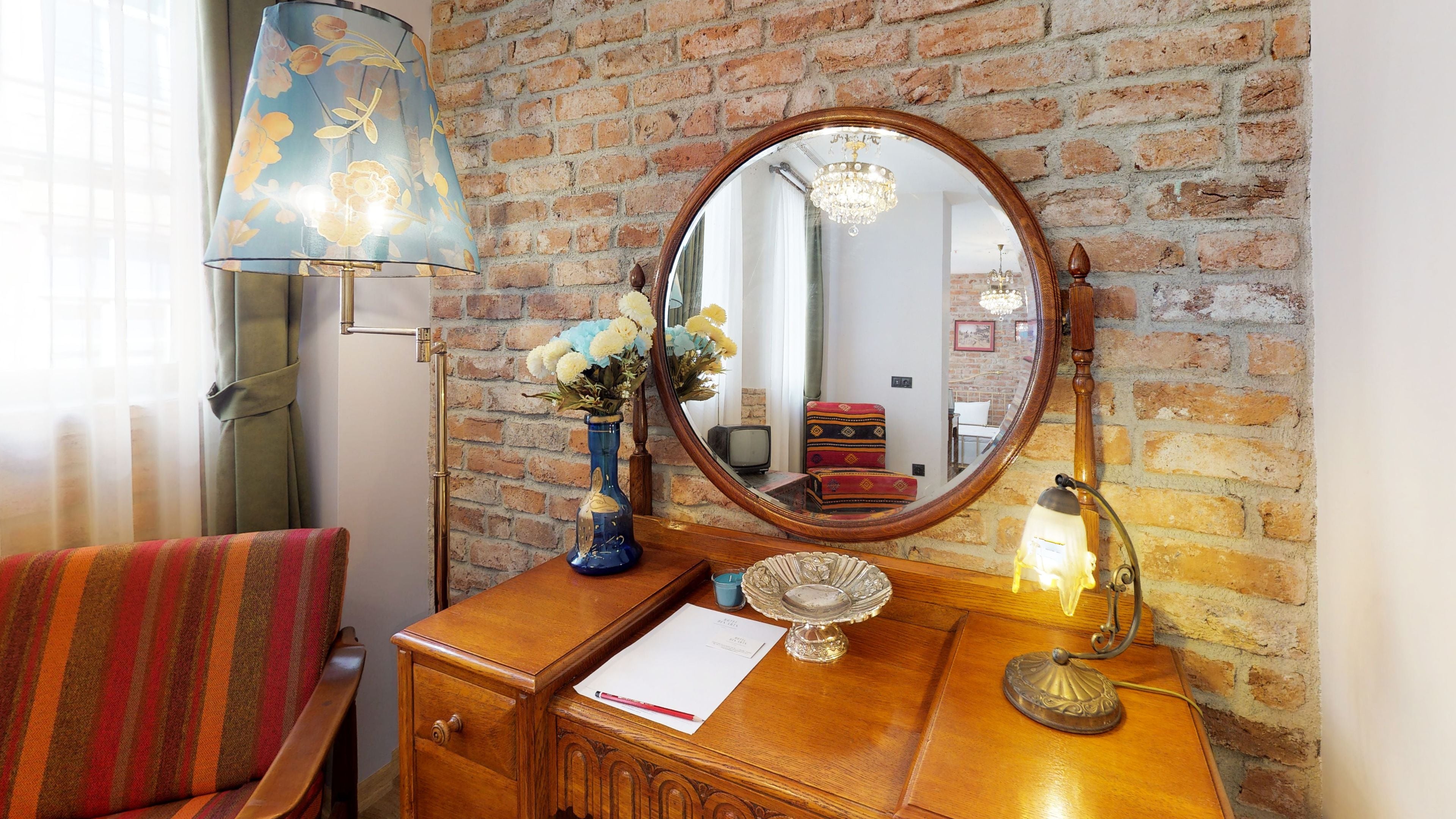 A wooden dresser with a mirror, a vase with flowers, a lamp, and a chair with a striped cushion.