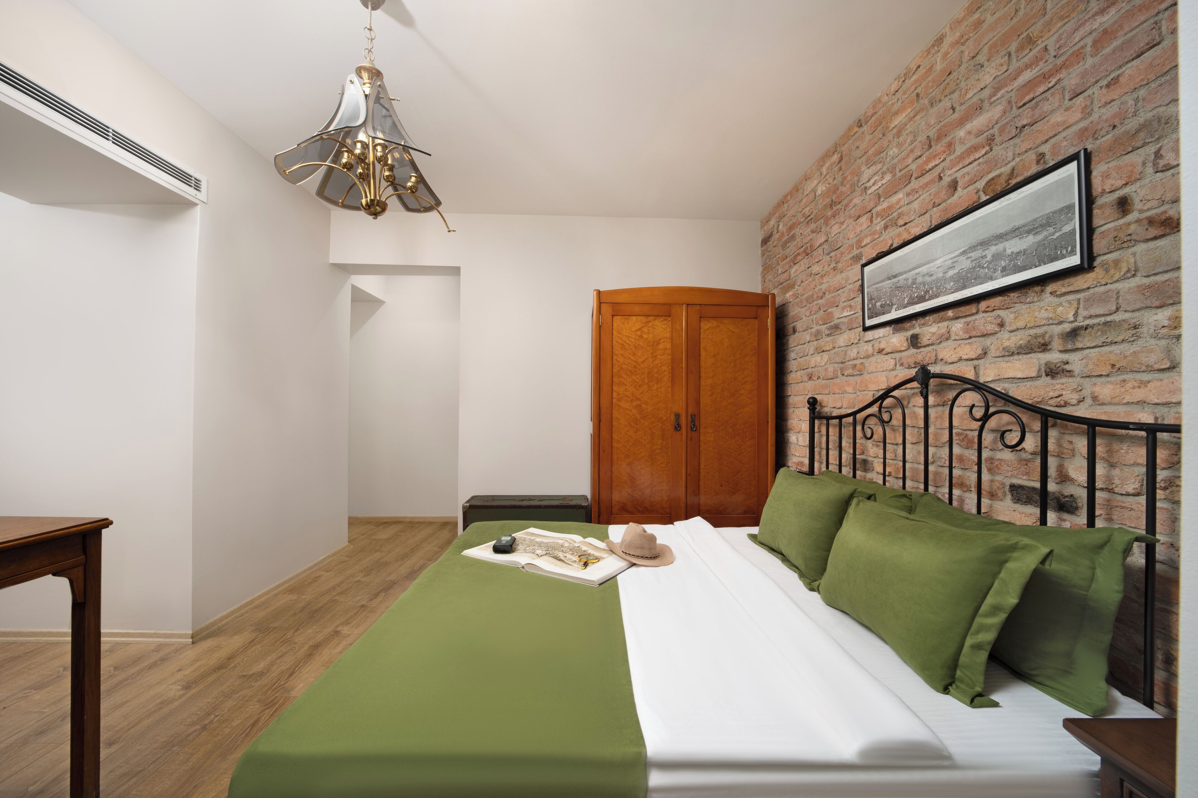 A bedroom with a brick wall, a wooden wardrobe, a bed with green and white bedding, and a hanging light fixture.
