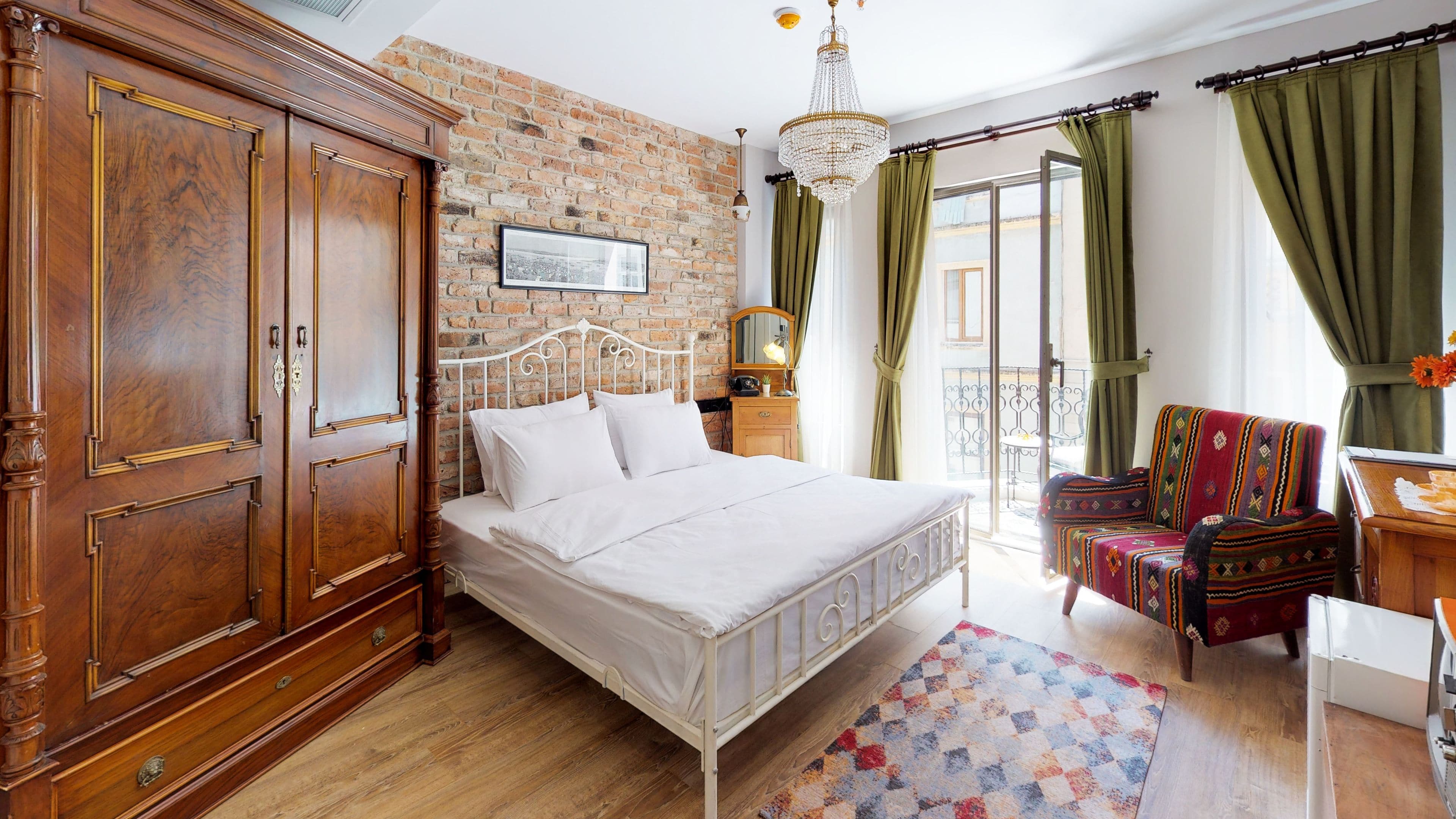 A bedroom with a brick wall, a large wooden wardrobe, a white metal bed frame, a colorful rug, and a balcony with green curtains.