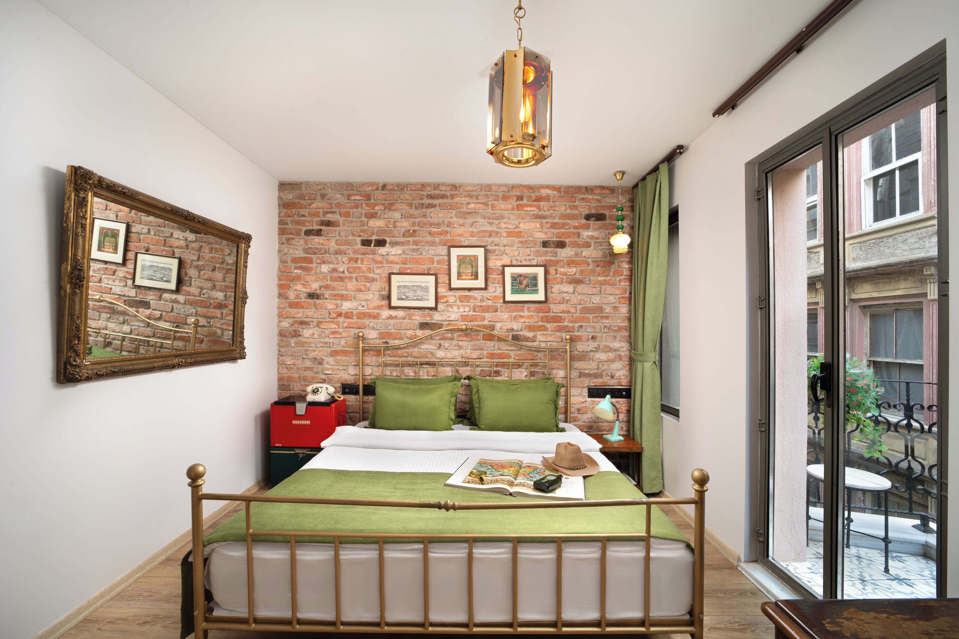 A room with a brick wall, a bed with green bedding, a hanging light fixture, and a window with a view of the outside.