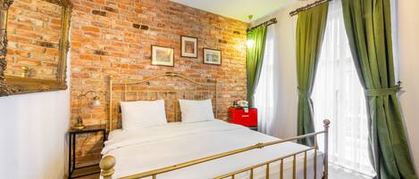 A room with a brick wall, a bed with a metal frame, a nightstand, a lamp, and a red box.