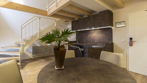 A modern kitchen with dark cabinets, a built-in oven, and a stainless steel range hood. There is a staircase with a white railing leading to an upper level. A potted plant is on a round table in the foreground. - Hotel Jakobslust (Gruenstadt)