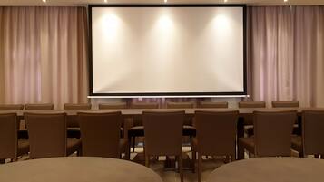 Un salón de conferencias con una pantalla grande, varias mesas y sillas.