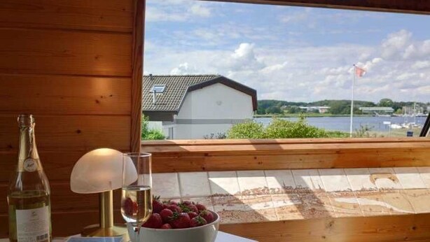 Ein Holzinterieur mit einem Tisch, auf dem eine Flasche, ein Glas und eine Schüssel mit Obst stehen, mit Blick auf ein Gewässer und Gebäude.