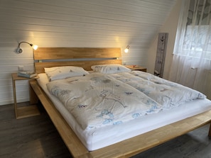 Una cama con estructura de madera y ropa de cama blanca con motivos náuticos, dos mesitas de noche con lámparas y una ventana con cortinas ligeras.