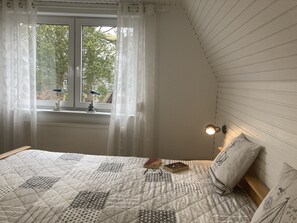Ein Schlafzimmer mit einem Bett, einem Fenster mit Vorhängen, einer Nachttischlampe und Blick auf Bäume draußen.