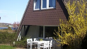 Ein Haus mit geneigtem Dach, einem kleinen Balkon mit weißen Stühlen und Blick auf ein Gewässer im Hintergrund.