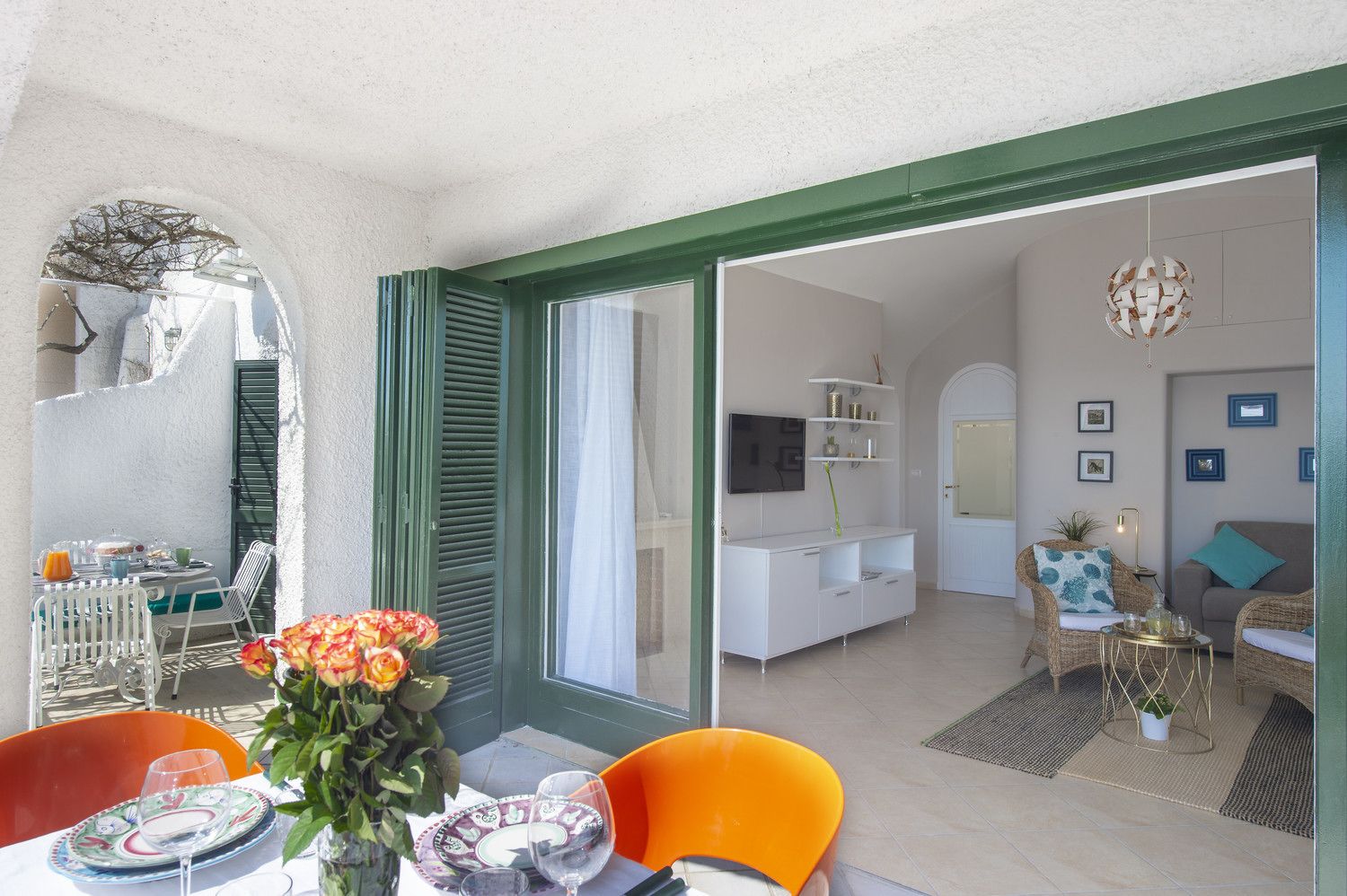 Un comedor con una mesa preparada para dos personas, un florero con flores y vistas a una sala de estar con televisor, estanterías y un sofá.