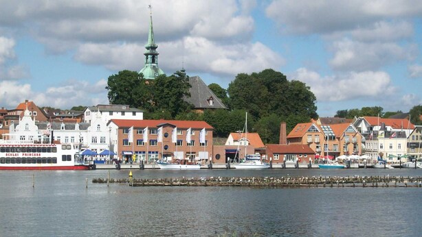 A waterfront town with a prominent church spire, a red-roofed building, and a white building with "SCHLEI PRINCESS" signage. - Apartment De lütte Stove - Apartment Woydt De lütte Stove (Kappeln)