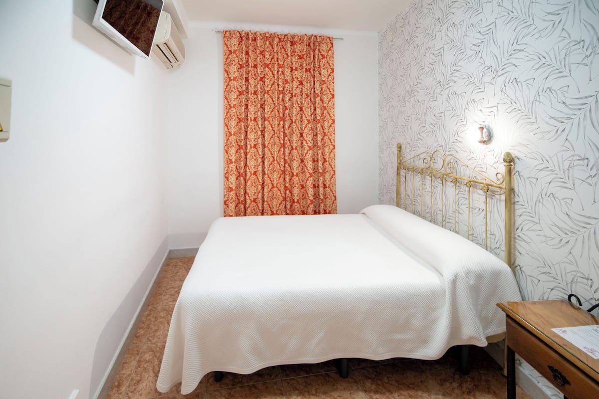 A small, neatly made bed with a white bedspread, a wooden nightstand with a lamp, and a patterned wall.