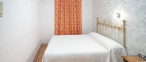A small, neatly made bed with a white bedspread, a wooden nightstand with a lamp, and a patterned wall.