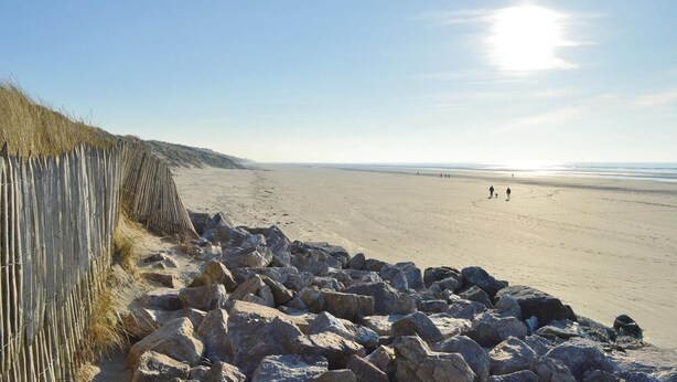Une plage avec une clôture en bois et de gros rochers au premier plan, et des personnes qui se promènent sur le sable à l’arrière-plan.